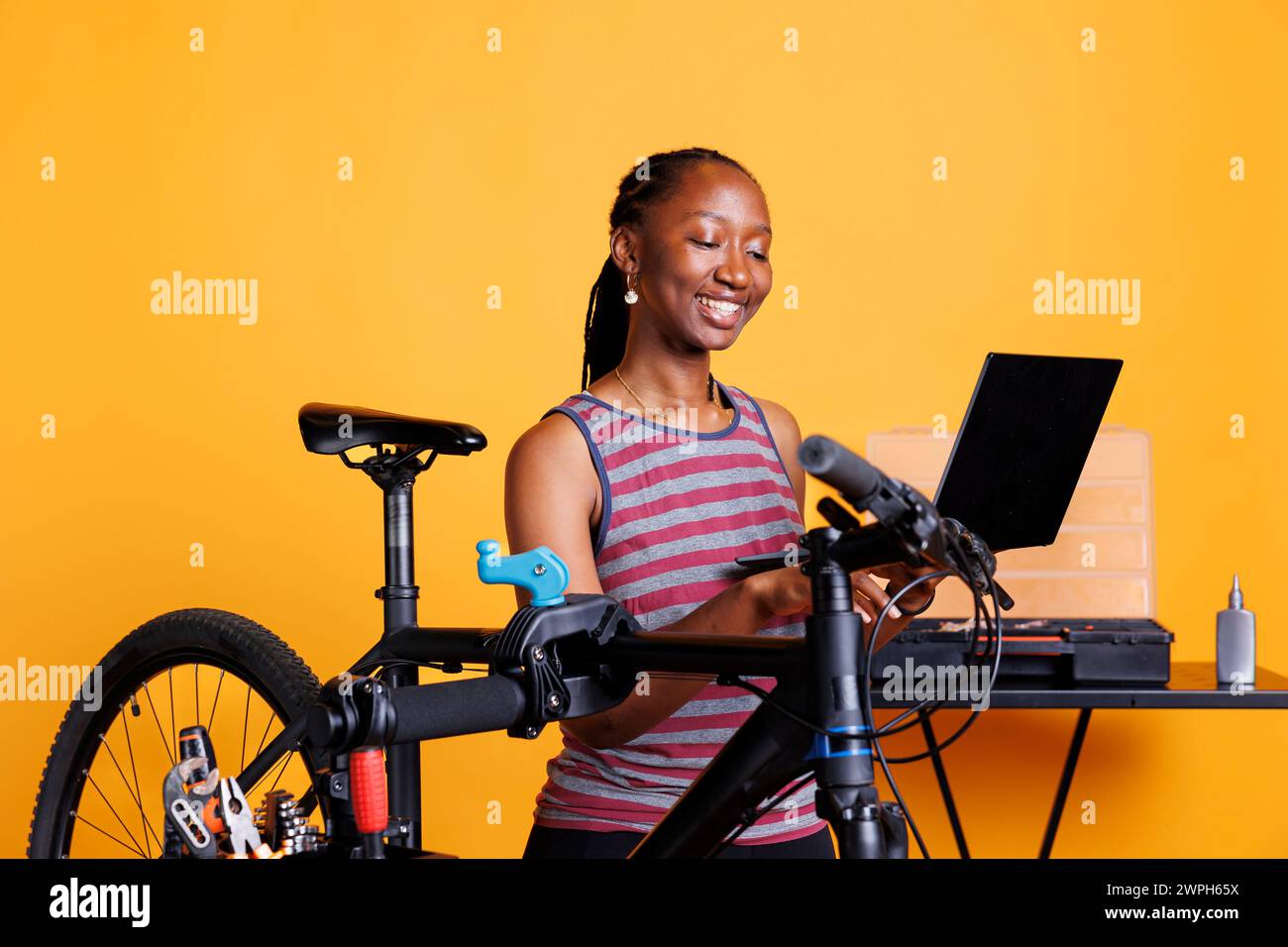 Energiegeladene schwarze Frau, die das Motorrad inspiziert und repariert, mit Laptop und speziellem Toolkit für Wartung und Reparaturen. Sportliche afroamerikanerin mit Minicomputer, die Fahrradschäden repariert. Stockfoto