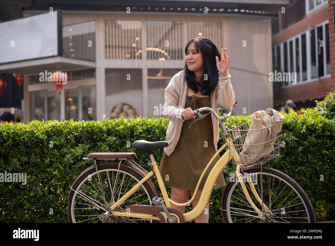 Eine attraktive, freundliche junge asiatische Frau in einem süßen Kleid winkt mit der Hand, um ihren Freund zu grüßen, während sie ihr Fahrrad in der Stadt schiebt. Eine Frau pendelt Stockfoto