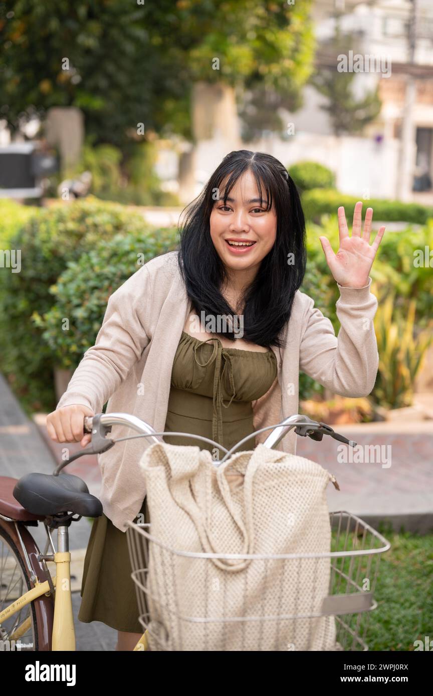 Eine attraktive, freundliche junge asiatische Frau in einem süßen Kleid winkt mit der Hand, um ihren Freund zu grüßen, während sie ihr Fahrrad in der Stadt schiebt. Eine Frau pendelt Stockfoto