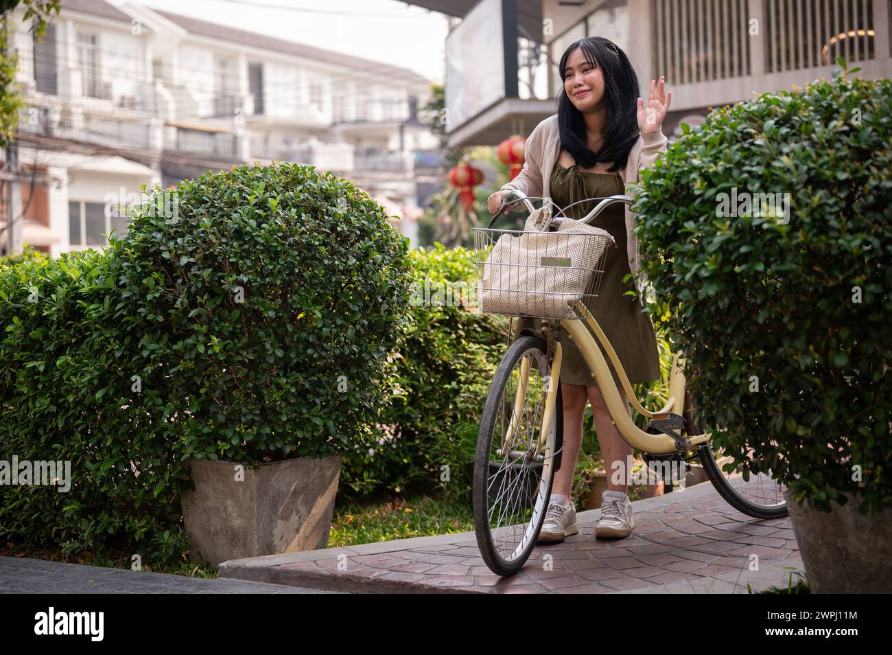 Eine attraktive, freundliche junge asiatische Frau in einem süßen Kleid winkt mit der Hand, um ihren Freund zu grüßen, während sie ihr Fahrrad in der Stadt schiebt. Eine Frau pendelt Stockfoto