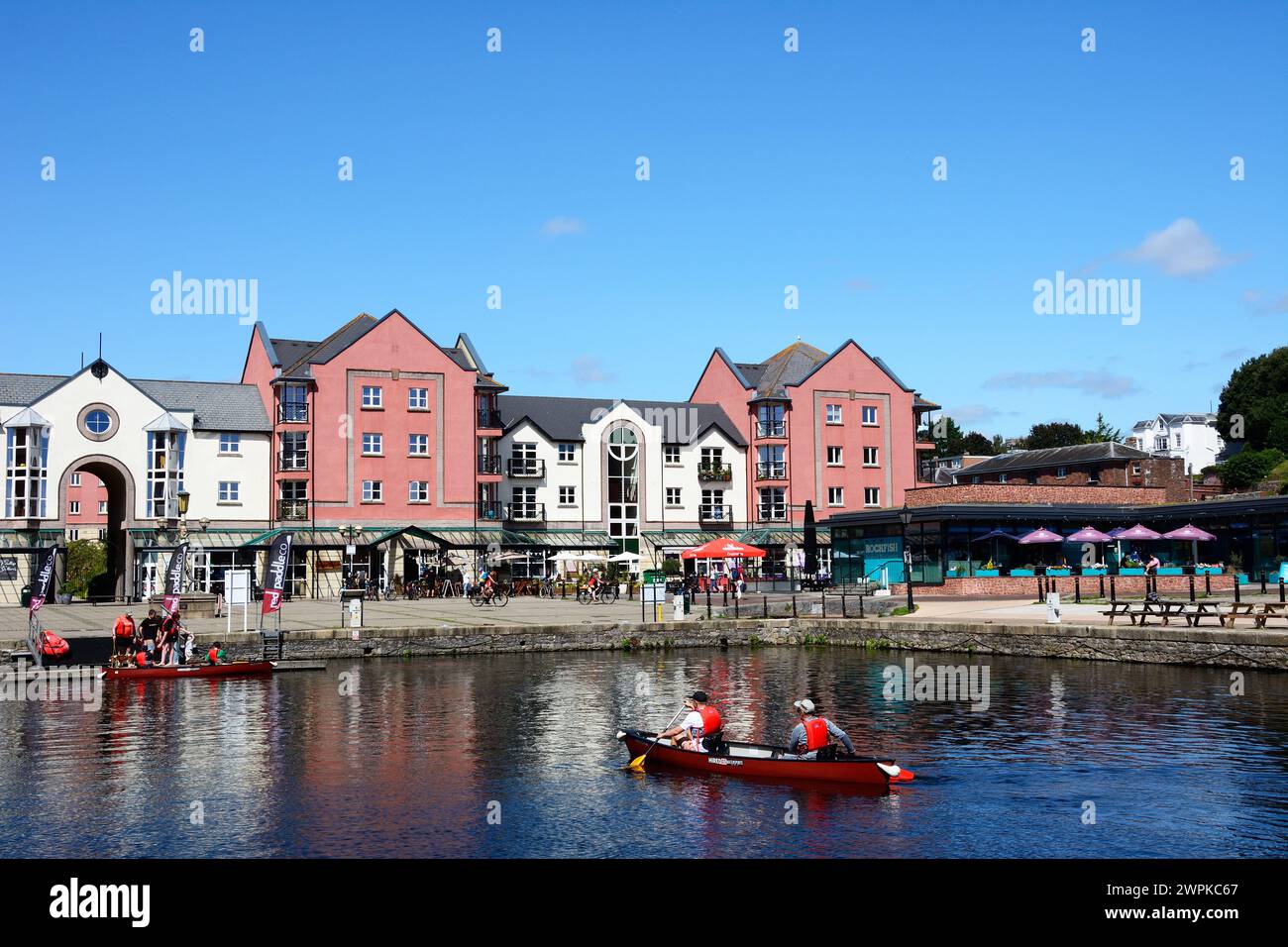 Menschen fahren Kajakfahren auf dem Exeter Schiffskanal mit Cafés und Restaurants auf der Piazza Terracina auf der Rückseite, Exeter, Devon, Europa. Stockfoto