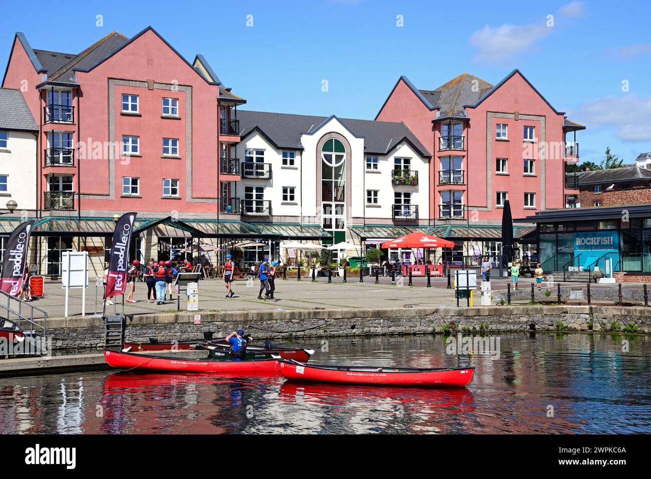Menschen fahren Kajakfahren auf dem Exeter Schiffskanal mit Cafés und Restaurants auf der Piazza Terracina auf der Rückseite, Exeter, Devon, Europa. Stockfoto