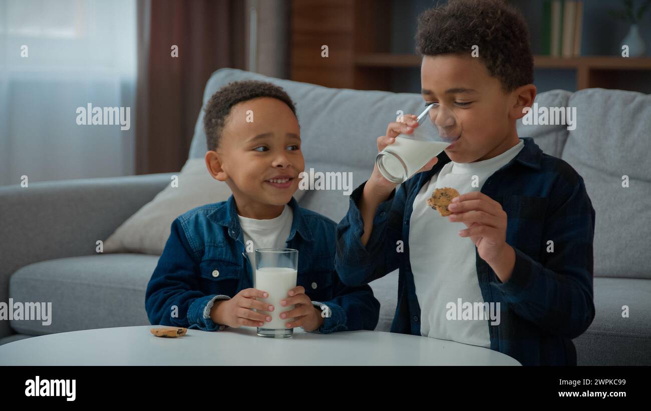 Zwei kleine afroamerikanische Jungen Kinder ethnisch adoptierte hungrige Kinder trinken Milch essen Kekse zu Hause Wohnzimmer Geschwister trinken Milchgetränk Stockfoto