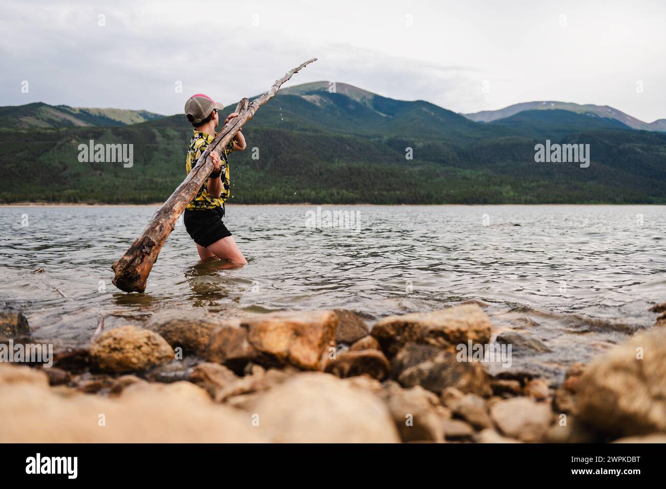 Junge, der mit einem großen Stock in einem ruhigen Bergsee spielt. Stockfoto