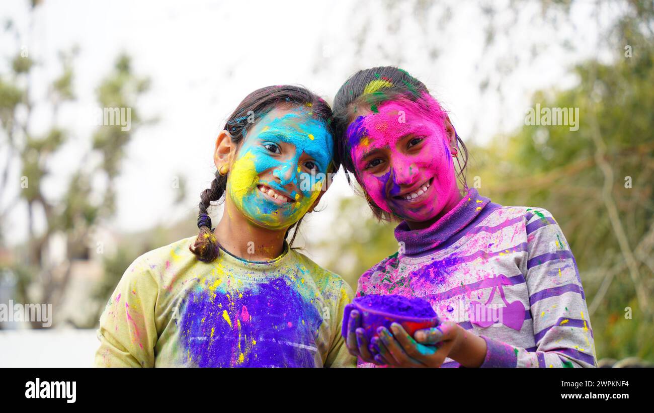 Glückliche indische Freunde oder Kinder, die das Hindufest von Holi feiern, indem sie sich gegenseitig Farben auftragen, beste Freunde. Weibliche Geschwister, die einen Teller halten Stockfoto