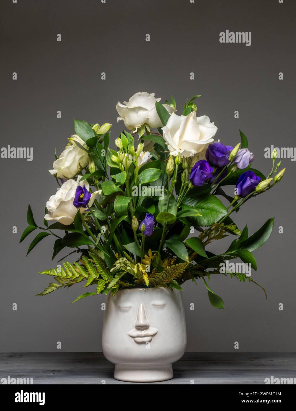 Blumenanordnung aus weißen Rosen, lila Lisianthus und Laub in einer weißen Keramik-Gesichtsvase. Stockfoto