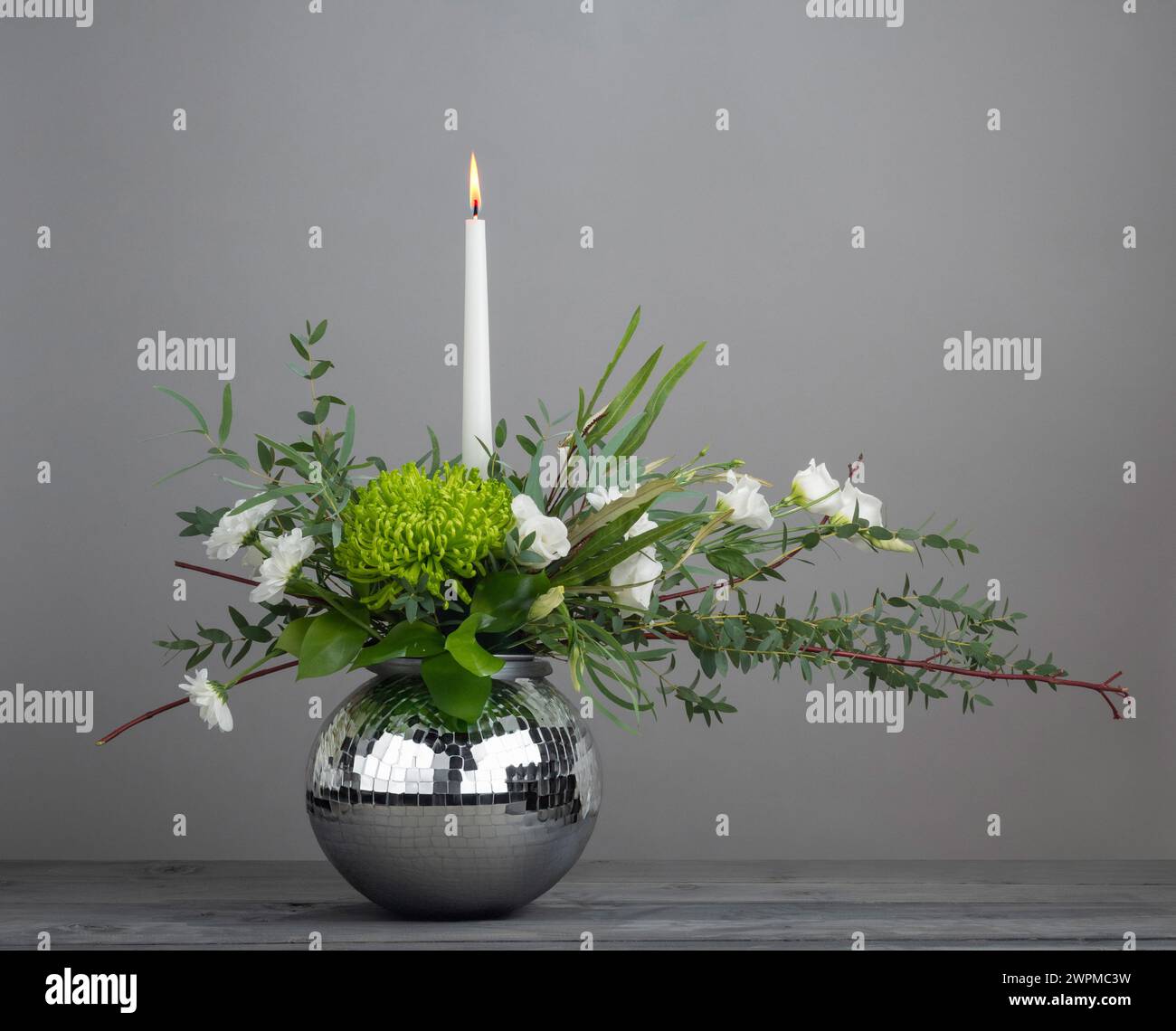 Blumenanordnung einer Kerze in einem kugelförmigen Spiegelkugelbehälter auf grauem Hintergrund. Stockfoto