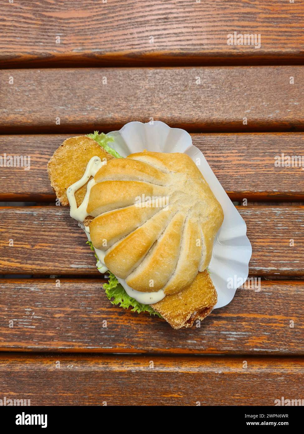 Ein Fischbrötchen in Form einer Muschel, belegt mit frittiertem Fisch und Tatarsauce in einem Karton auf einem Holztisch Stockfoto