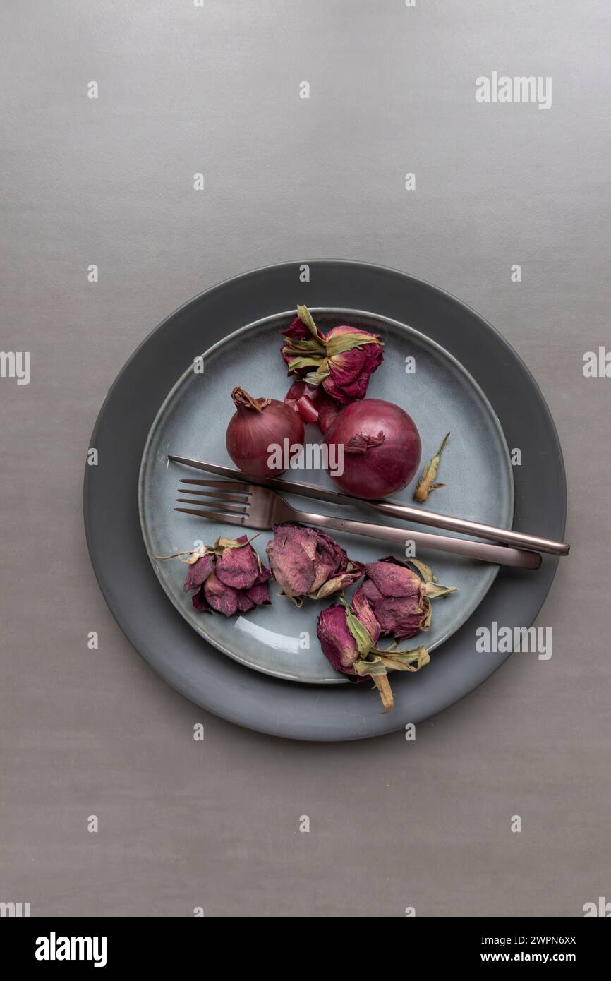 Anordnung von zwei ganzen roten Zwiebeln mit getrockneten Rosenblättern neben kupferfarbenem Besteck auf einer Porzellanplatte als Stillleben Stockfoto