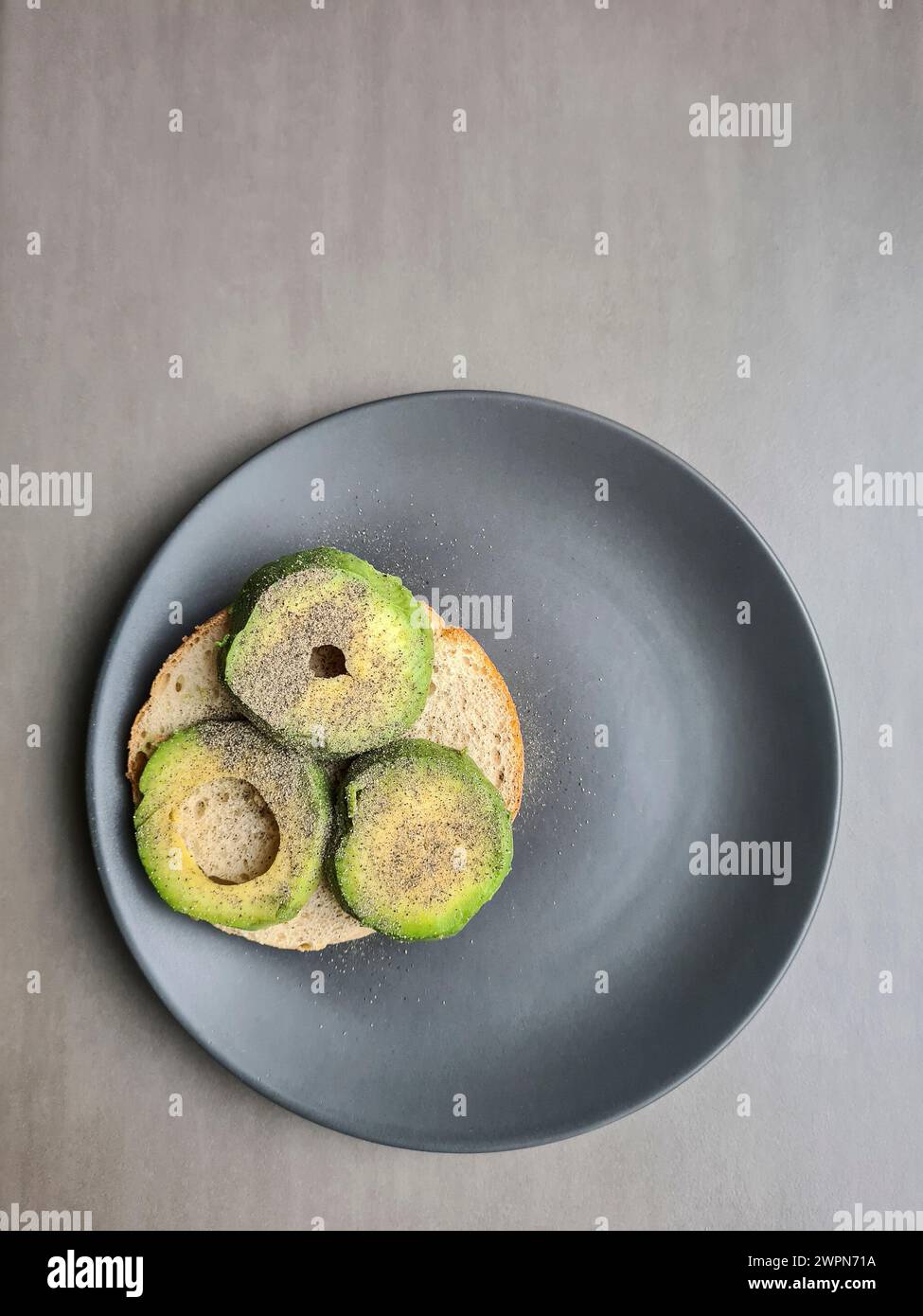 Eine in Scheiben geschnittene Avocado, die mit Pfeffer gewürzt wurde, liegt auf einem halben Brötchen auf einem grauen Teller vor grauem Hintergrund Stockfoto