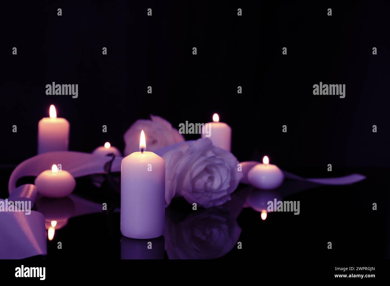 Violette Rosen und brennende Kerzen auf schwarzer Spiegelfläche in Dunkelheit, Raum für Text. Beerdigungsattribute Stockfoto