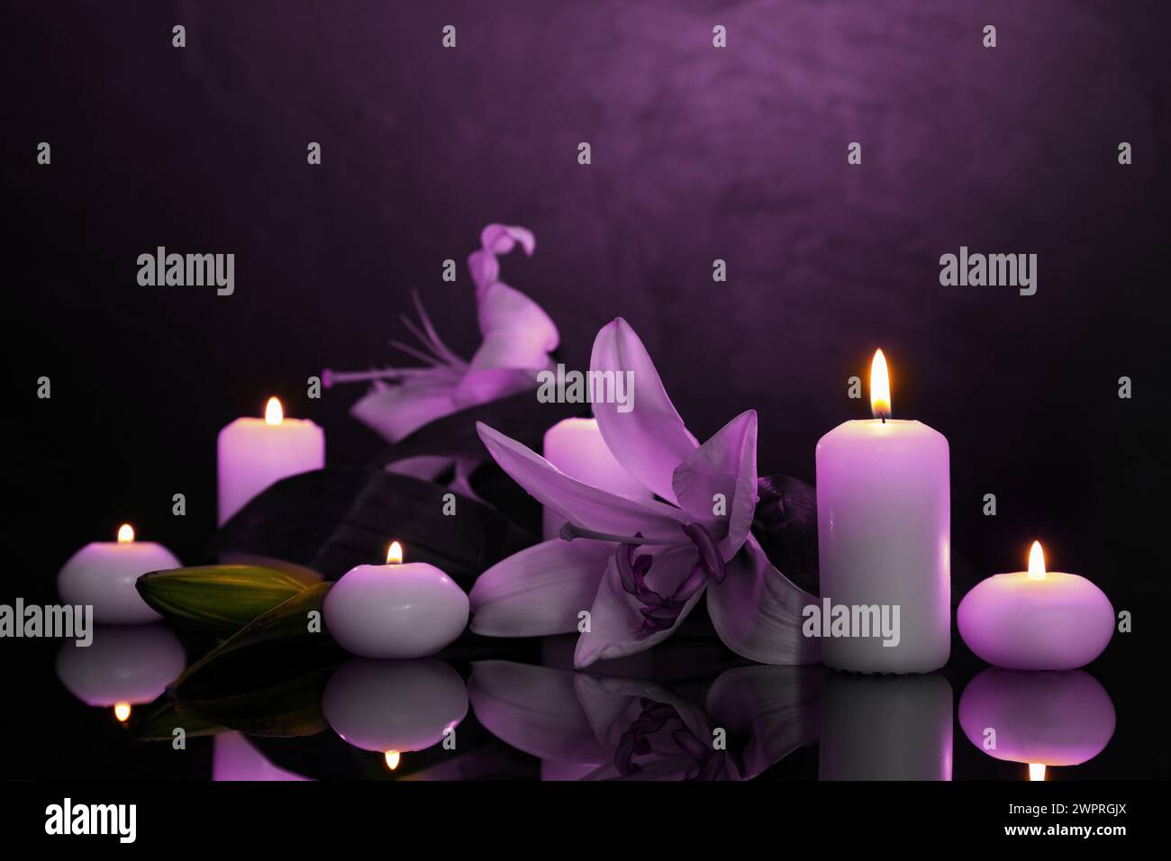 Violette Lilien und brennende Kerzen auf schwarzer Spiegeloberfläche in der Dunkelheit. Beerdigungsattribute Stockfoto