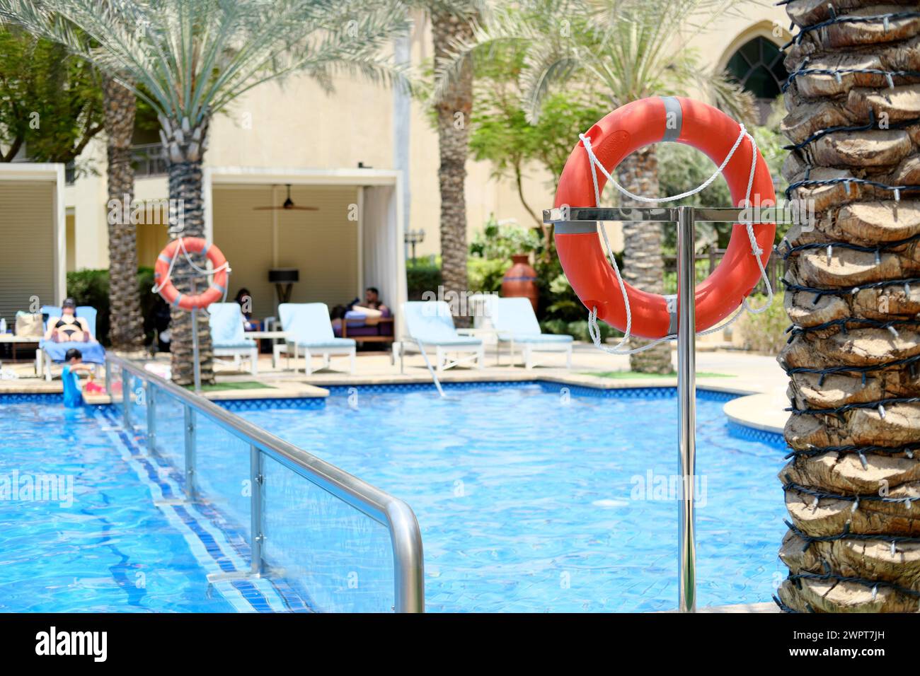 Ein leuchtend roter Rettungsschirm liegt vor dem ruhigen blauen Wasser des Palace Downtown Dubai Pools, in dem Gäste im Hintergrund entspannen. Dubai, VAE - August Stockfoto