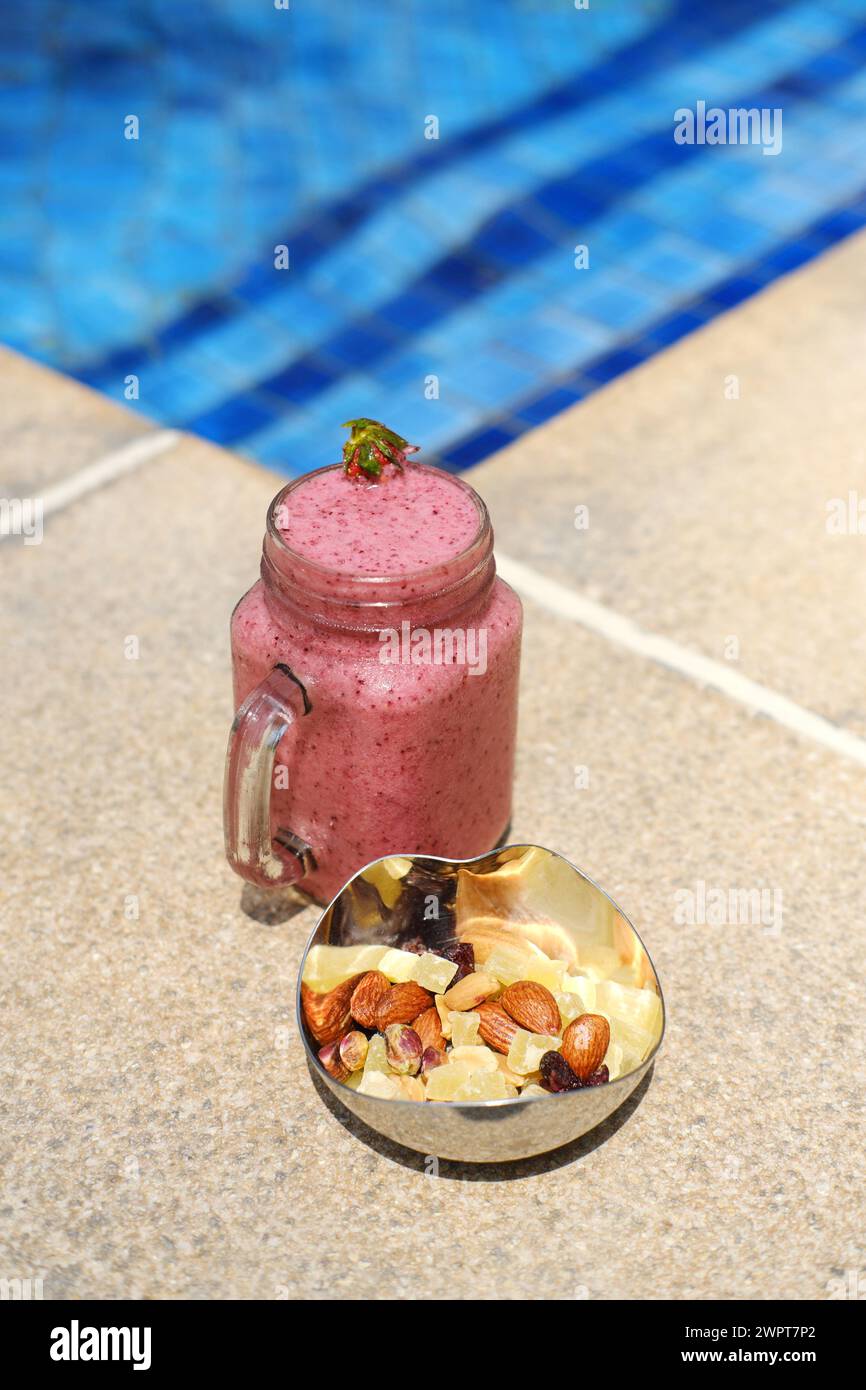 Ein heißer rosafarbener Beeren-Smoothie in einem Glas und eine Schüssel gemischter Nüsse neben einem glitzernden blauen Swimmingpool. Vertikales Foto Stockfoto