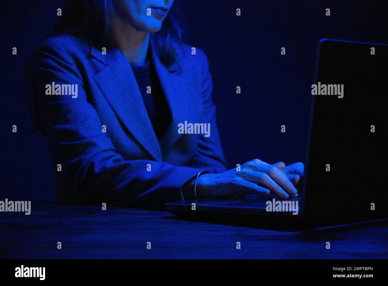 Geschäftsfrau, die im Dunkeln an einem Laptop arbeitet und vom Bildschirm beleuchtet wird. Keine Fläche. Konzentrieren Sie sich auf die Hände. Blaue Farbkorrektur. Stockfoto