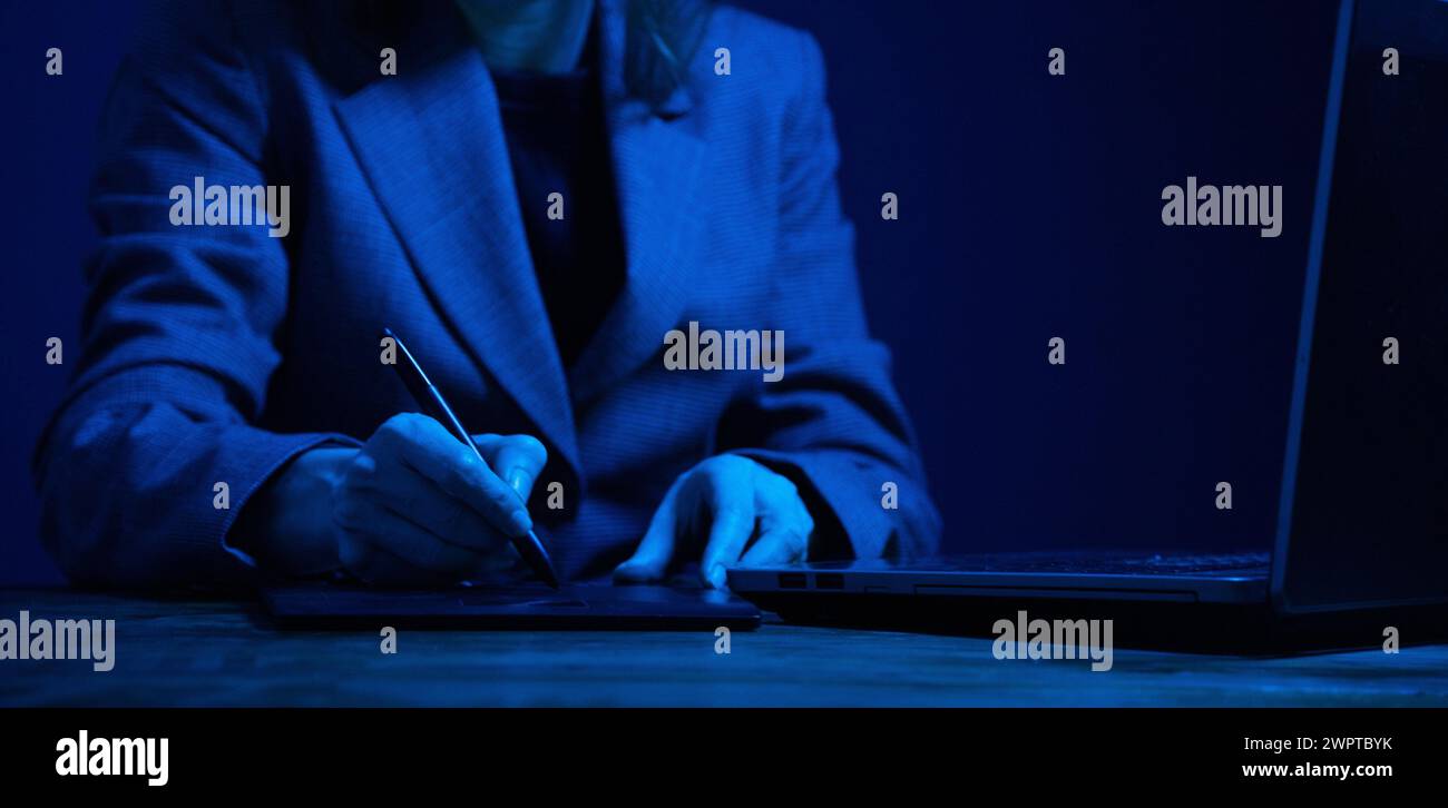 Geschäftsfrau, die im Dunkeln an einem Grafiktablett und Laptop arbeitet und vom Bildschirm beleuchtet wird. Keine Fläche. Konzentrieren Sie sich auf die Hände. Graufarbkorrektur. Stockfoto