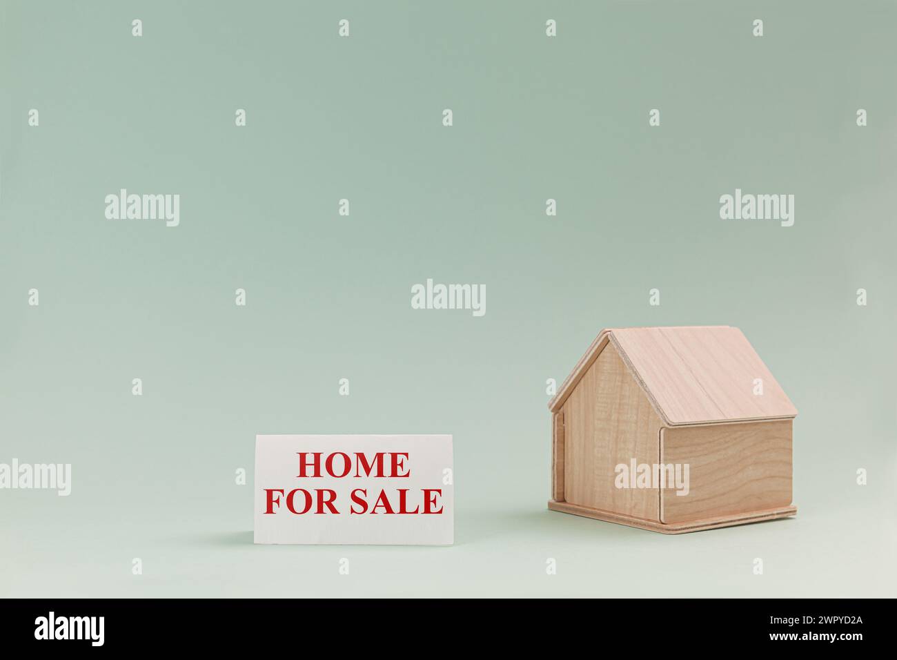 Vereinfachtes hölzernes Hausmodell isoliert auf blassgrünem Hintergrund, mit Text Haus zum Verkauf auf dem Schild. Stockfoto