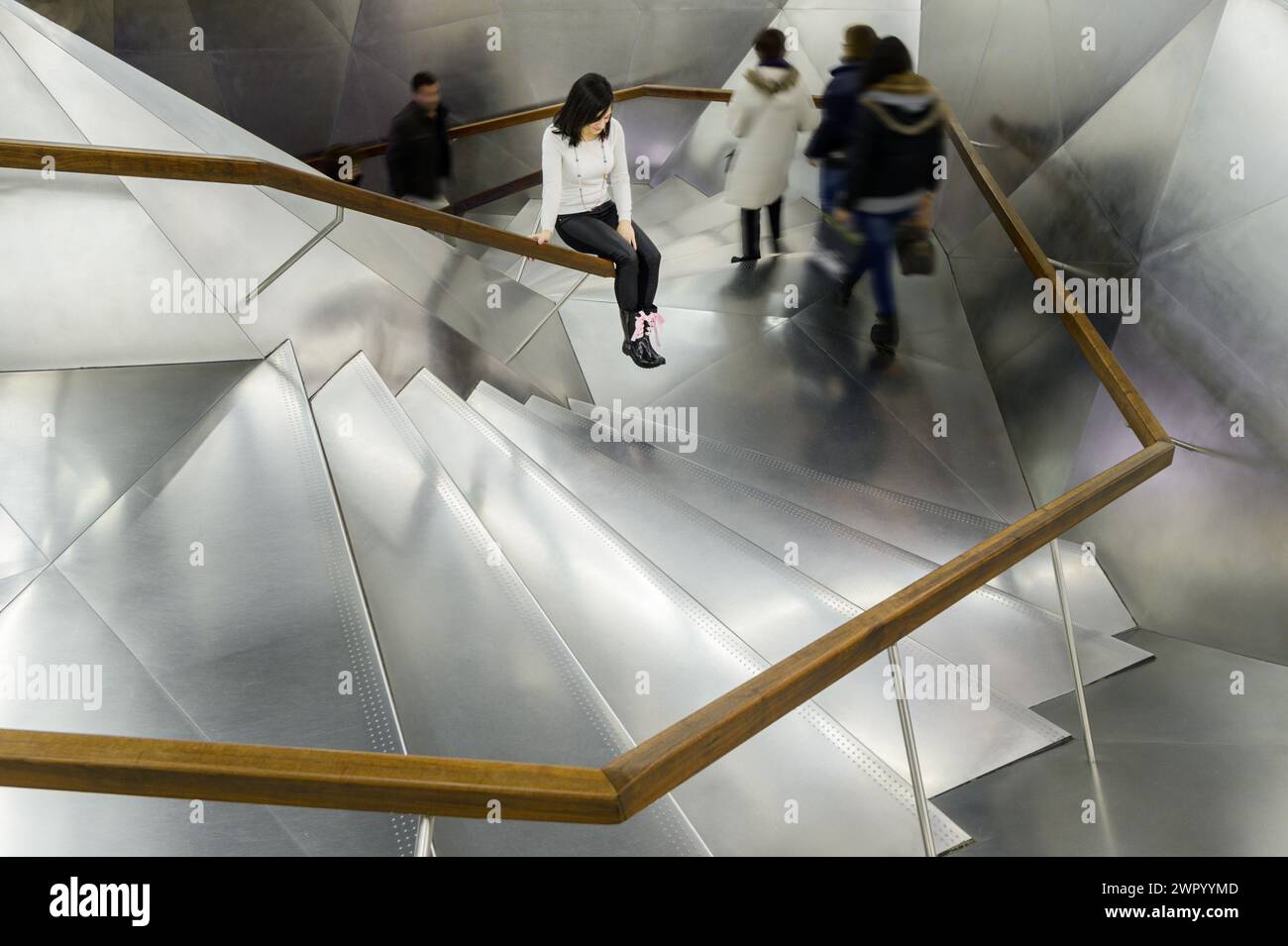 Ein Mädchen, das auf einem Geländer aus moderner Aluminiumtreppe sitzt, während die Menschen hoch und runter laufen, in Madrid, Spanien. Stockfoto