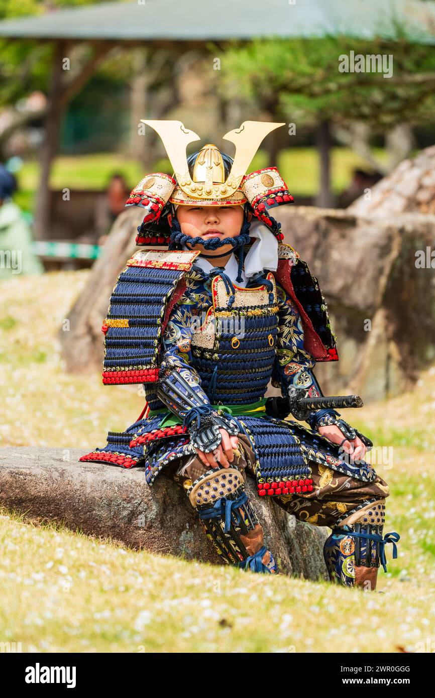 Junge, japanisches Kind, in Samurai-Rüstung gekleidet, sitzt auf einem großen Stein auf der Burg Tatsuno während der Samurai-Parade im Frühjahr. Augenkontakt. Stockfoto