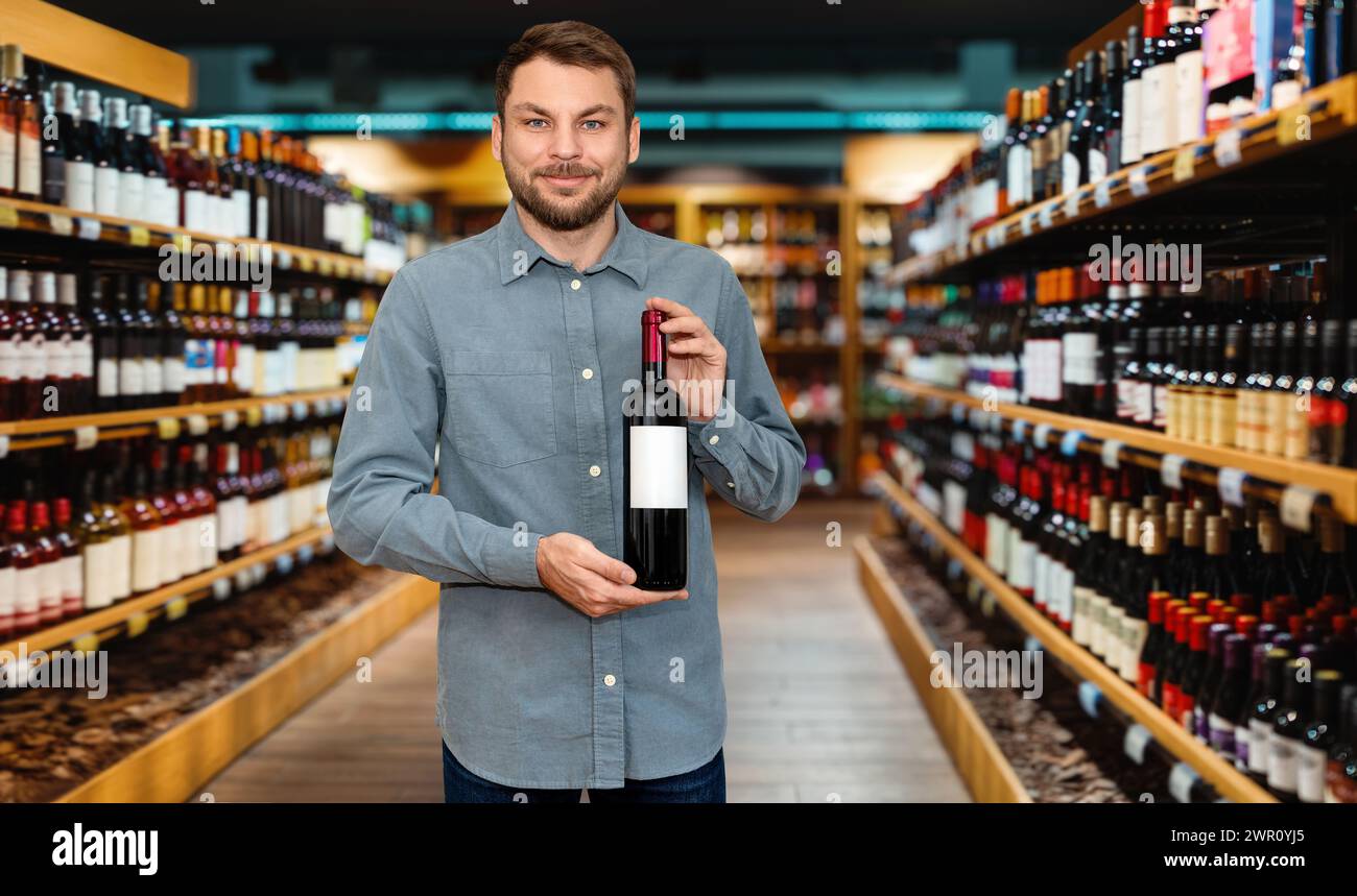 Fröhlicher Kundenmann, der eine Flasche Wein in der Hand hält und im Spirituosenladen lächelnd. Stockfoto