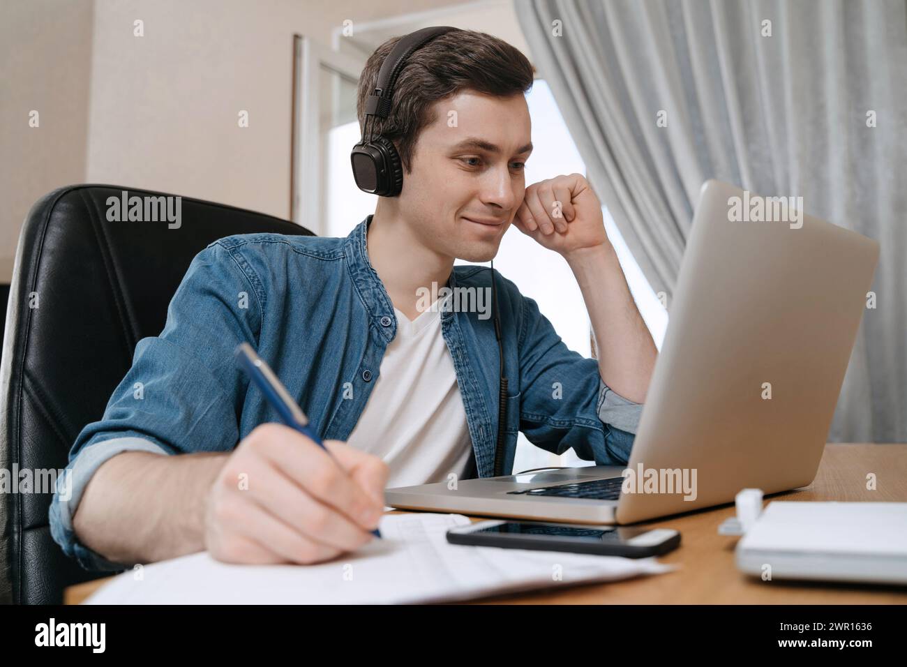Glücklicher Schüler, der Kopfhörer mit Laptop für Fernunterricht trägt, Videokonferenzen genießt, Webinar ansieht, Audiokurse anhört, Notizen schreibt, mit dem Lehrer lernt, Lektion haben Stockfoto