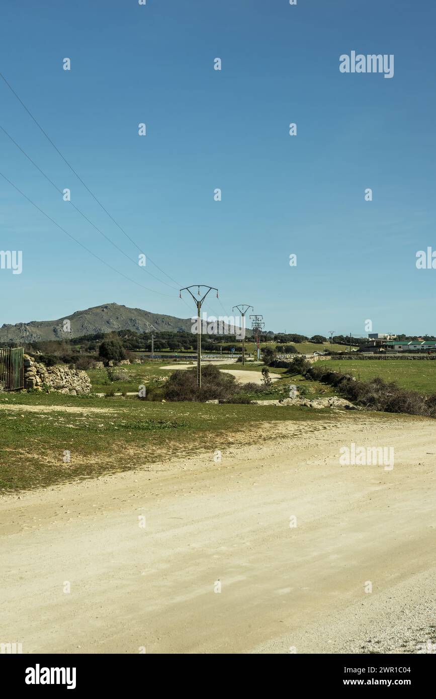 Ein ländlicher Feldweg, flankiert von Grasfeldern und einer Stromleitung auf Hochmasten Stockfoto