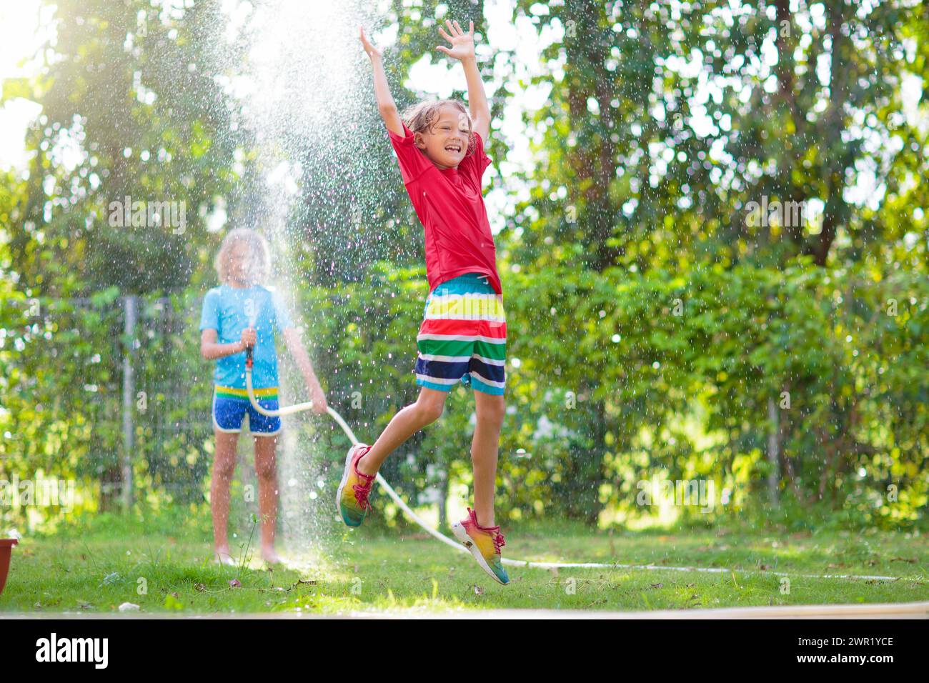 Kinder spielen mit einem Wasserschlauch. Sommergarten Outdoor Spaß für Kinder. Ein Junge spritzt Wasser an einem heißen, sonnigen Tag. Kinder, die Pflanzen im Garten bewässern. Stockfoto
