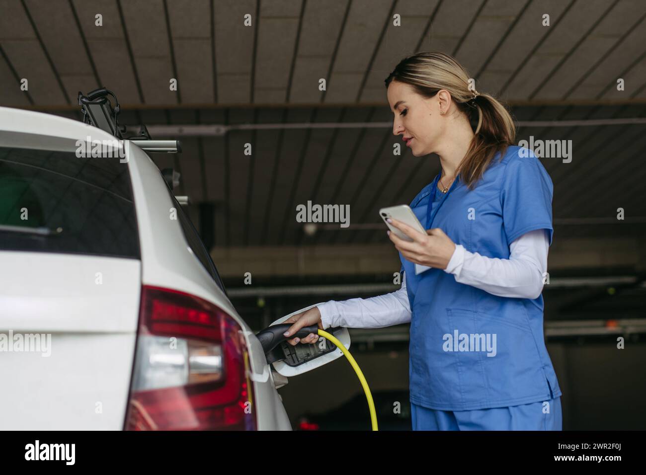 Das Ladegerät wird vor der Arbeit von der Schwester an das Elektroauto angeschlossen. Elektrofahrzeug-Ladestation vor dem Krankenhaus, moderne Klinik. Laden am Arbeitsplatz Stockfoto