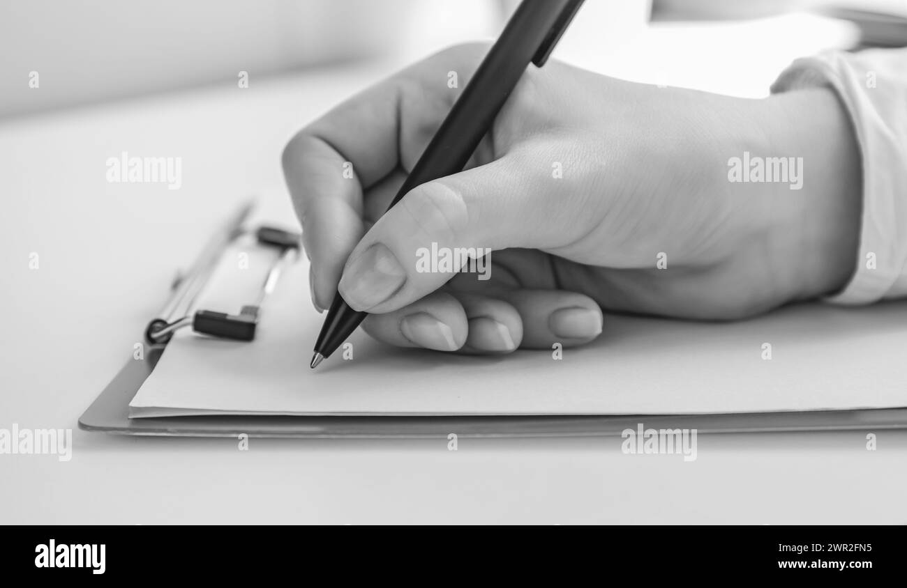 Schwarzweißfoto. Konzentriere dich auf eine weibliche Hand, die einen Stift hält, um etwas auf ein leeres Blatt Papier zu schreiben. Stockfoto