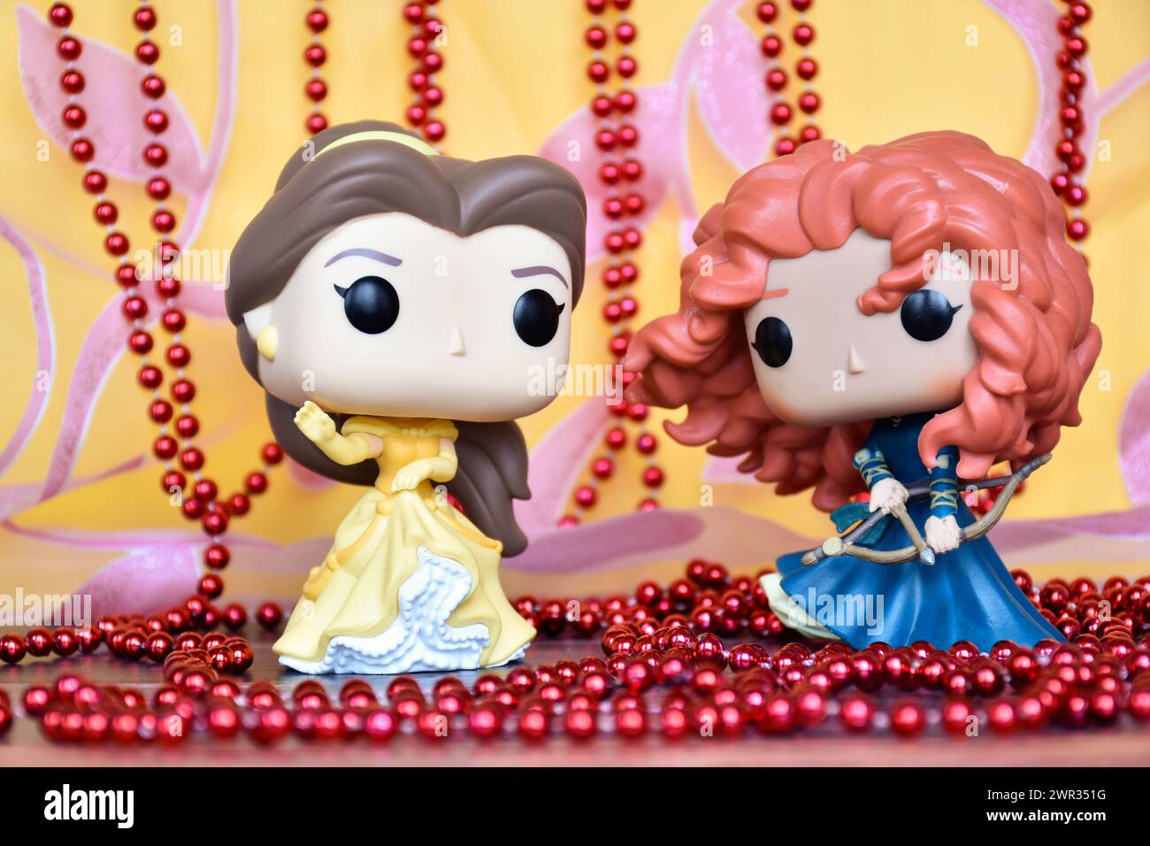 Funko Pop Actionfiguren der Disney Prinzessinnen Belle (die Schönheit und das Biest) und Merida (die Tapferkeit). Goldener rosa Vorhang, rote Halskette, fabelhafter Palast. Stockfoto