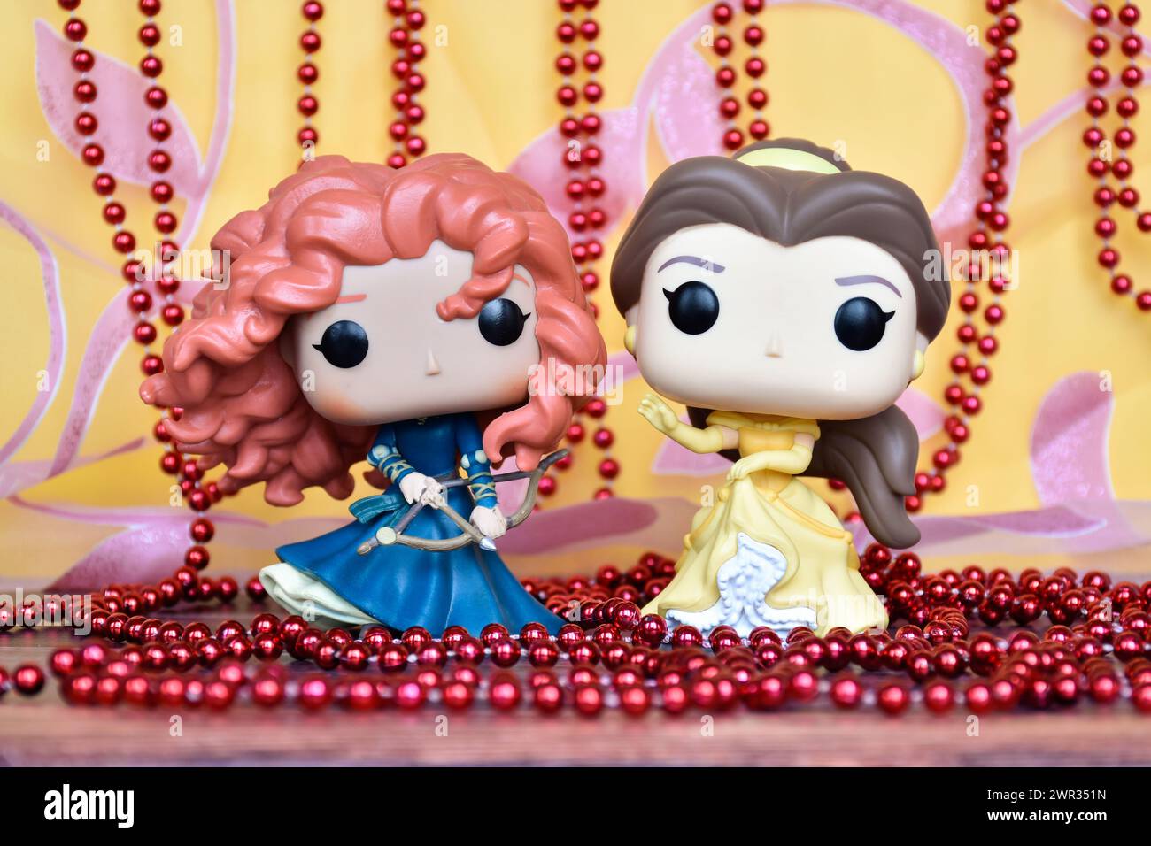 Funko Pop Actionfiguren der Disney Prinzessinnen Merida (tapfer) und Belle (Schönheit und das Biest). Goldener rosa Vorhang, rote Halskette, fabelhafter Palast. Stockfoto