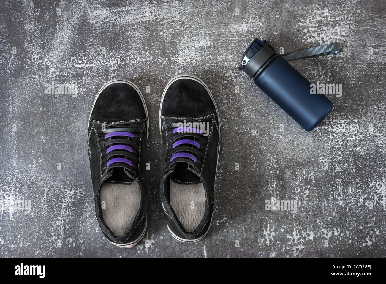 Schwarze Sportschuhe mit zweifarbigen Schnürsenkeln und einem blauen Kunststoffbehälter mit Wasser auf grauer Oberfläche Stockfoto