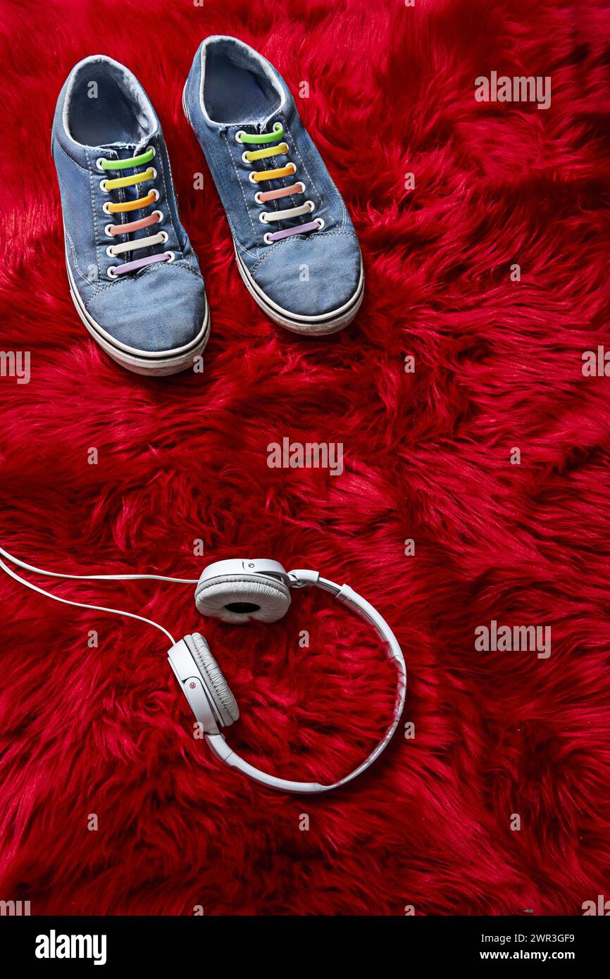 Blaue Sneakers mit bunten Schnürsenkeln und weißen Kopfhörern mit Kabelanschluss auf roter Haaroberfläche Stockfoto
