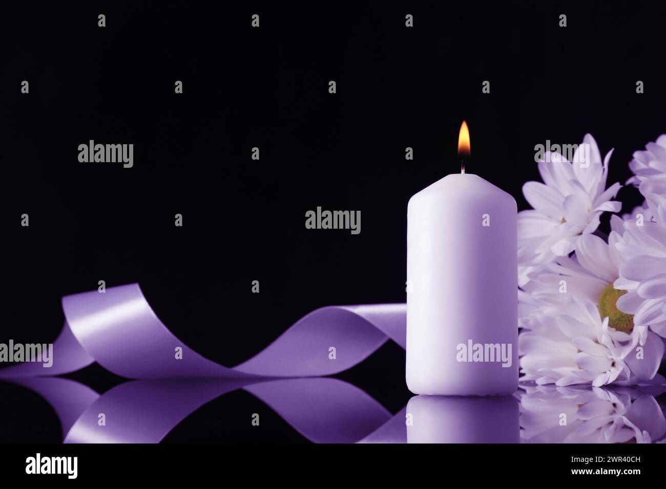 Brennende Kerze, violette Chrysanthemenblumen und Band auf schwarzer Spiegelfläche in Dunkelheit, Raum für Text. Beerdigungsattribute Stockfoto