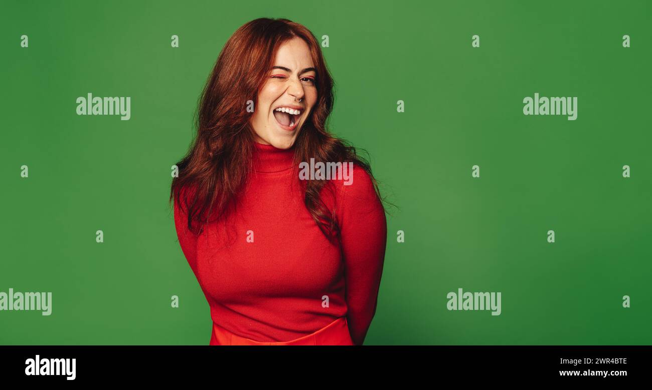Modische Frau mit Septumring steht in einem Studio mit grünem Hintergrund. Sie zwinkert und lächelt glücklich. Ihre trendige Freizeitkleidung und ihre lebendige Statur Stockfoto