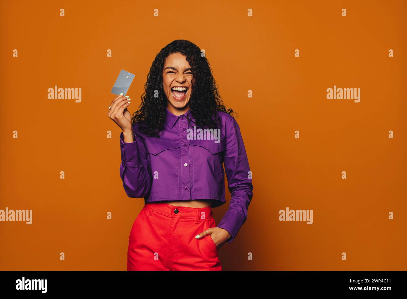 Frau in lässiger Kleidung steht vor einem orangen Hintergrund, hält eine Karte und strahlt vor Aufregung. Sie zahlt mühelos mit kontaktlosem Tippen Stockfoto