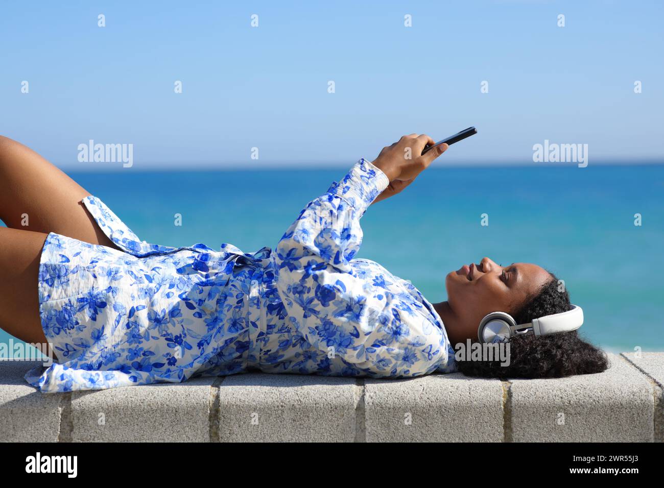 Profil einer glücklichen schwarzen Frau, die Kopfhörer trägt und Medien am Telefon ansieht, die am Strand liegen Stockfoto