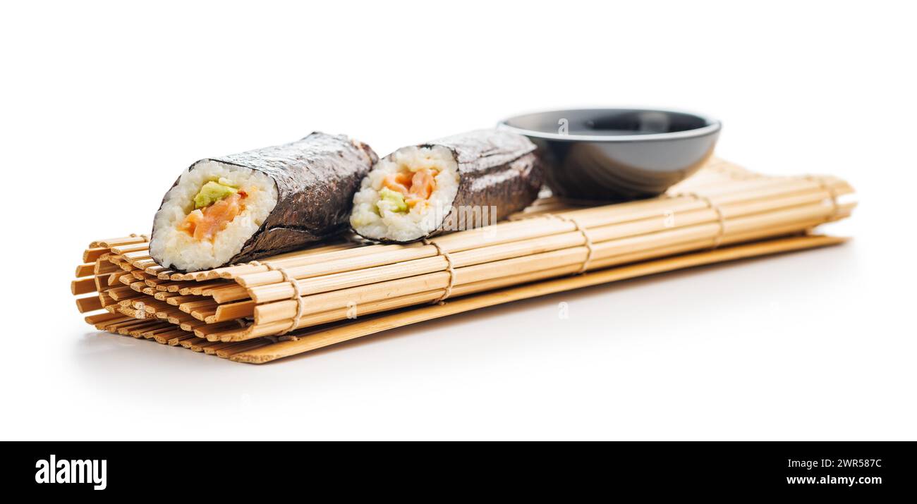 Eine Bambusmatte mit zwei Sushi-Brötchen, die ordentlich neben einer Schüssel Sojasauce platziert werden. Die Sushi-Brötchen sind mit Reis, Algen und verschiedenen Zutaten gefüllt. Stockfoto