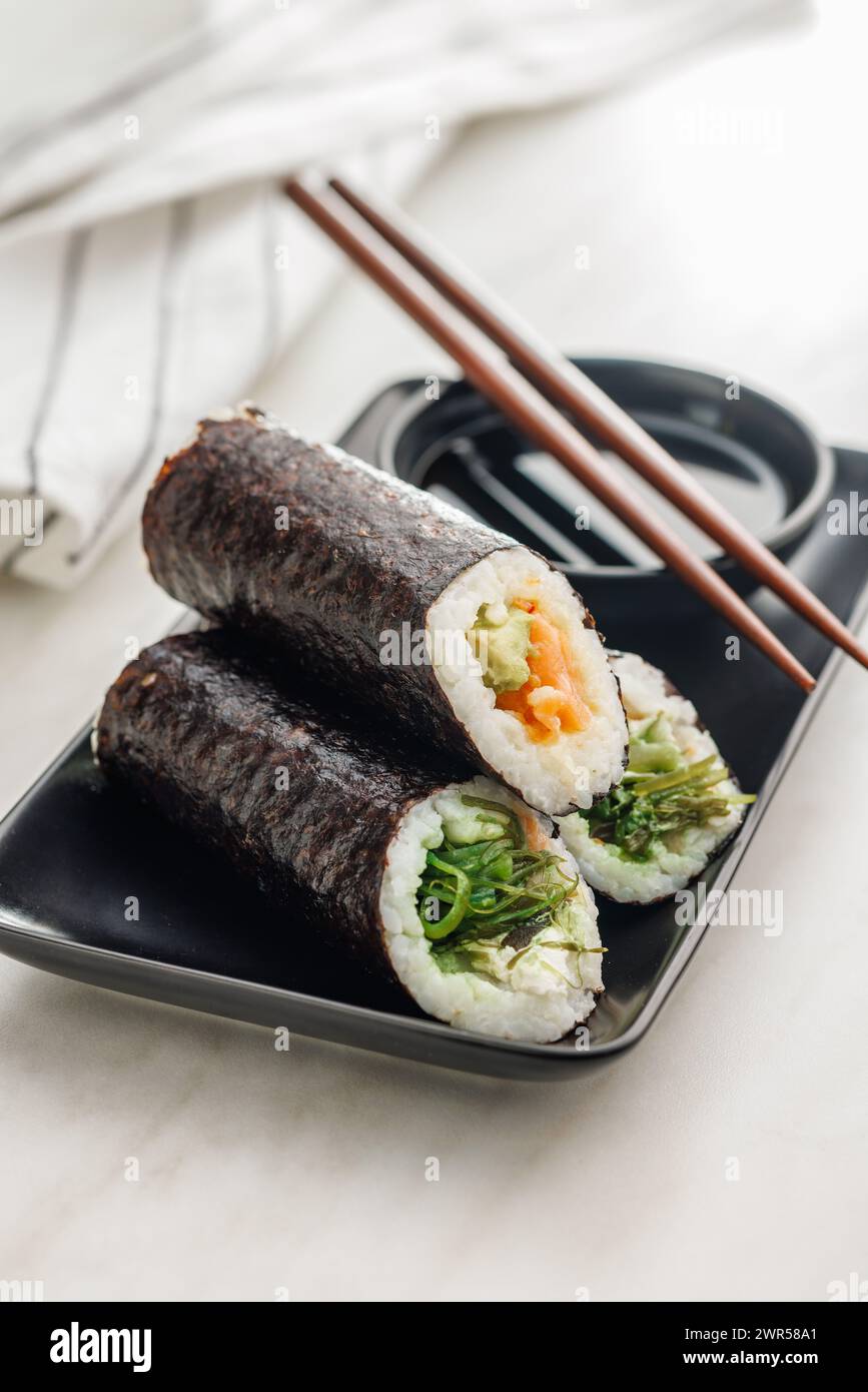 Ein Teller gefüllt mit verschiedenen Sushi-Brötchen mit einem Satz Holzstäbchen und einer Schüssel Sojasauce auf einem einfachen Küchentisch. Stockfoto