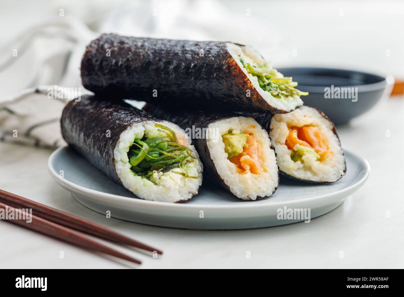 Ein Teller gefüllt mit verschiedenen Sushi-Brötchen mit einem Satz Holzstäbchen und einer Schüssel Sojasauce auf einem einfachen Küchentisch. Stockfoto