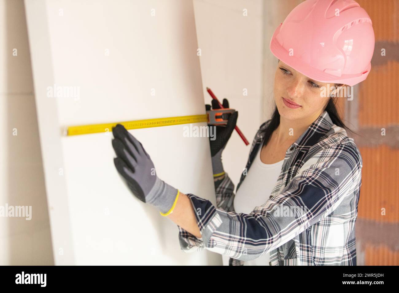 Junge Bauarbeiterin mit rosa Schutzhelm, die die Wand misst Stockfoto