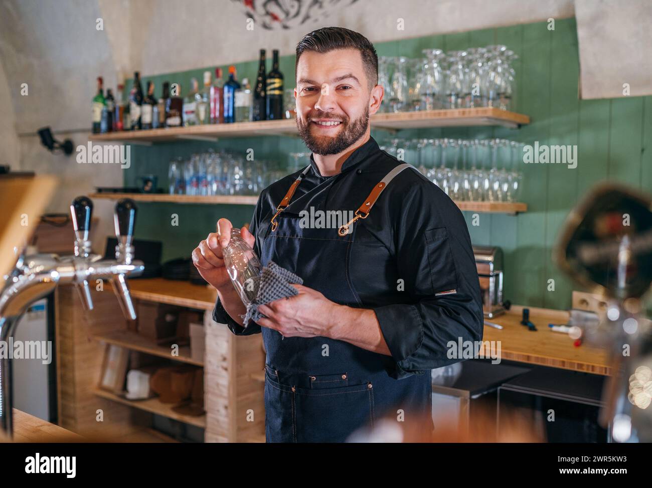 Porträt eines fröhlich lächelnden bärtigen Barkepers in schwarzer Uniform mit Schürze, der das Bierglas an der Bar abwischt. Erfolgreiche Mitarbeiter, harte Arbeit, Stockfoto