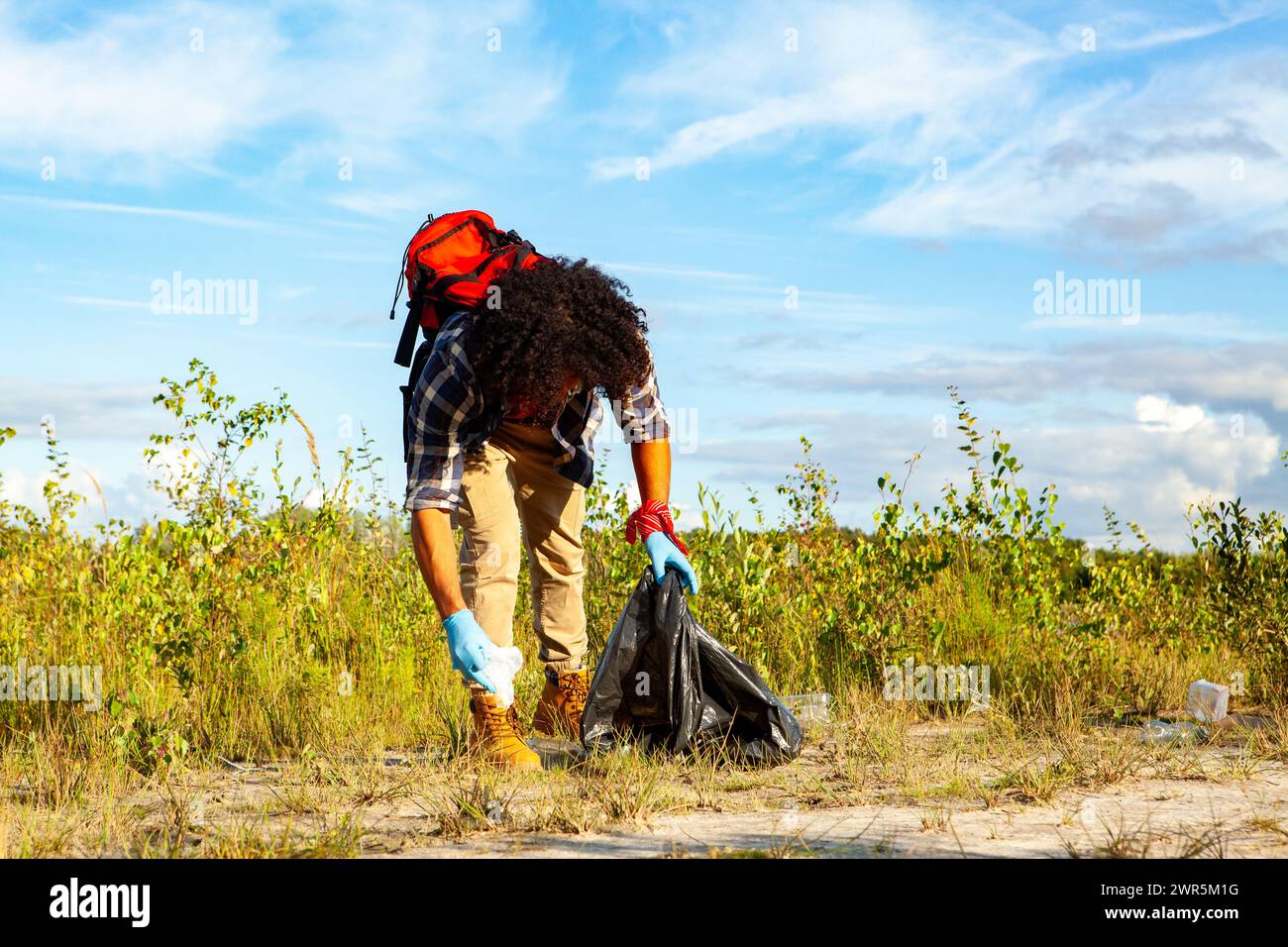 Eine Person mit lockigen Haaren, die Handschuhe trägt, sammelt Müll in einer schwarzen Tasche in einer natürlichen Umgebung und trägt so zum Umweltschutz unter einem klaren blauen Himmel bei. Freiwilliger Reinigungsstreu auf einem Feld an einem sonnigen Tag. Hochwertige Fotos Stockfoto