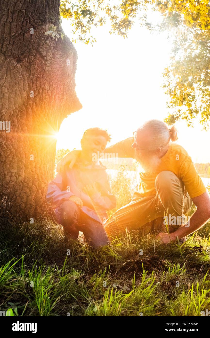 Dieses eindrucksvolle Bild fängt eine herzerwärmende Interaktion zwischen einem Großvater und seinem Enkel im sanften, goldenen Licht der untergehenden Sonne ein. Der ältere Mann, mit weißem Bart und gelbem Hemd, hockt sich in die Hocke, um auf Augenhöhe mit dem kleinen Kind zu sein, das ebenfalls hockt. Sie sind an einem großen Baum, und die Sonne erstrahlt durch die Äste und umgibt sie mit einer warmen, leuchtenden Atmosphäre. Der Großvater scheint Weisheit oder eine Geschichte mit dem Kind zu teilen, das aufmerksam zuhört. Es ist ein Bild von Generationenbindung und den einfachen, kostbaren Momenten, die das Gefüge meiner Familie bilden Stockfoto