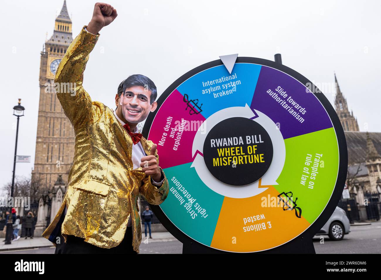 London, Großbritannien. März 2024. Ein Schauspieler, der eine Rishi Sunak Maske trägt und als Gastgeber einer Gameshow gekleidet ist, steht neben einem „Rad des Unglücks“ auf dem Parlamentsplatz während einer Veranstaltung, die von Amnesty International UK inszeniert wurde, um die schwerwiegenden negativen Folgen des Gesetzes zur Sicherheit Ruandas der Regierung hervorzuheben. Das House of Lords wird am 12. März über das umstrittene Gesetz abstimmen, und Amnesty International UK fordert Peers auf, das Gesetz abzulehnen. Quelle: Mark Kerrison/Alamy Live News Stockfoto
