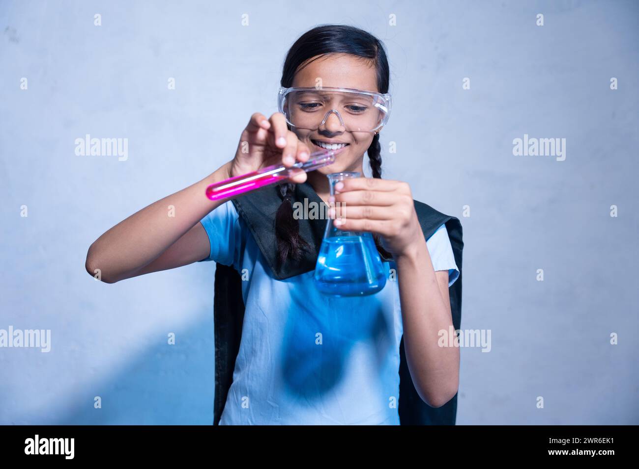 Glückliches junges Indianermädchen in Schuluniform, das eine Laborbrille trägt, während es an ihrem Chemieprojekt arbeitet, Experiment mit Reagenzgläsern und konischen flas Stockfoto