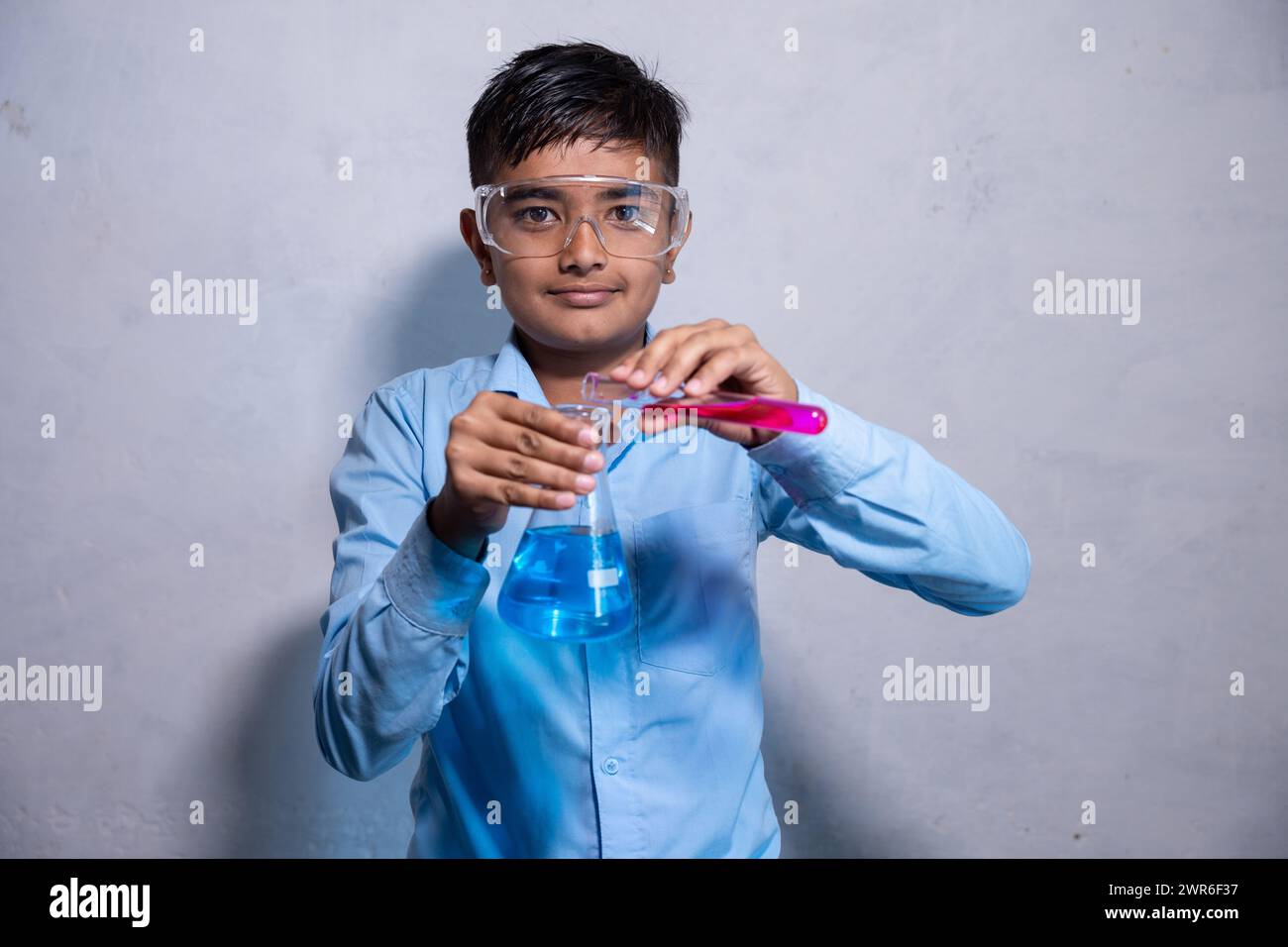 Glücklicher junger indianischer Junge in Schuluniform mit einer Sicherheitsbrille während der Arbeit an seinem Chemieprojekt Experiment mit Reagenzglas und konischem Kolben Stockfoto