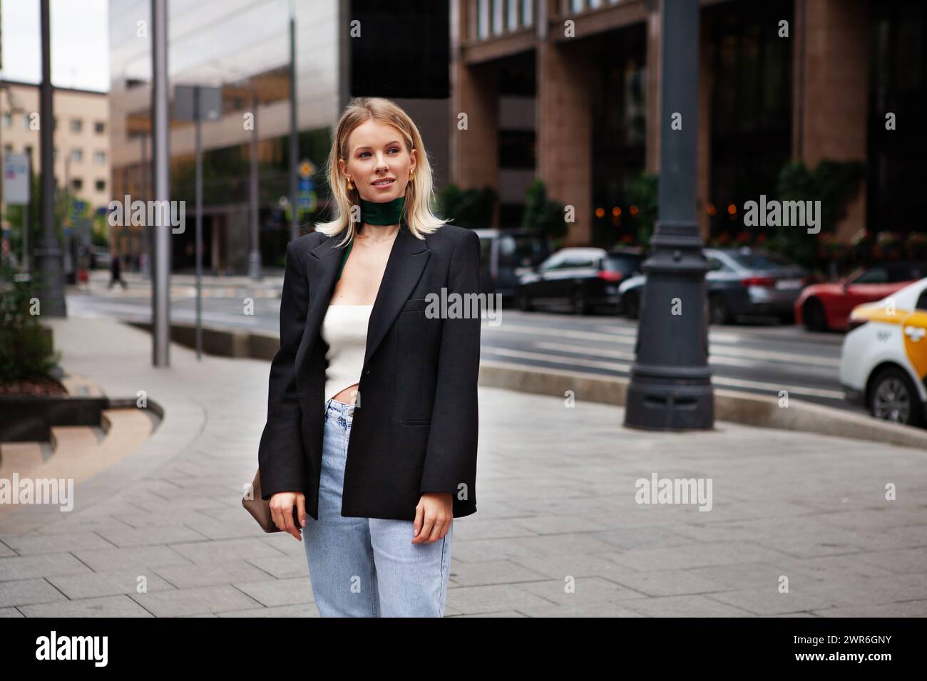 Schöne junge, stilvolle blonde Frau auf der Stadtstraße vor dem Hintergrund des Gebäudes, mit modischer Kleidung, schwarzem Blazer und Seidenschal. Modische Mo Stockfoto
