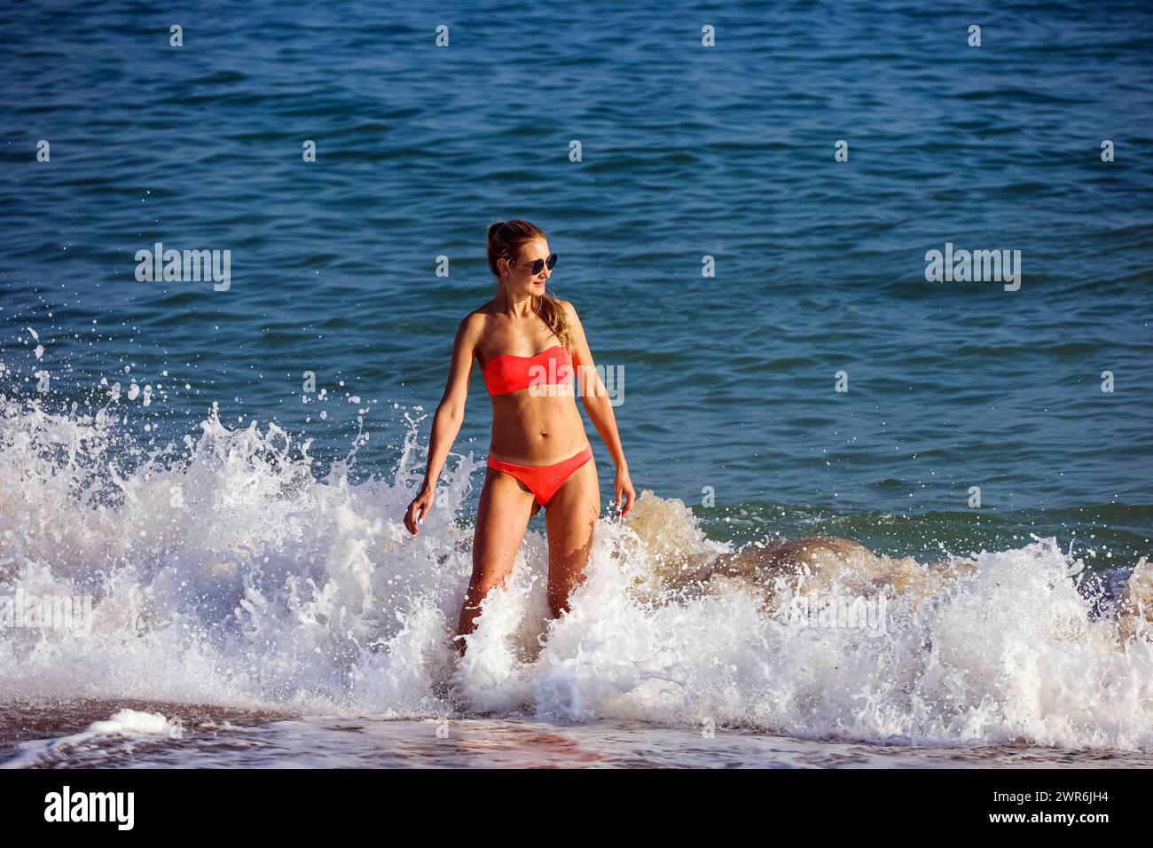Junge Frau in rotem Bikini und Sonnenbrille genießt Wasserspritzer am Strand in der Yalong Bucht der Insel Hainan Stockfoto