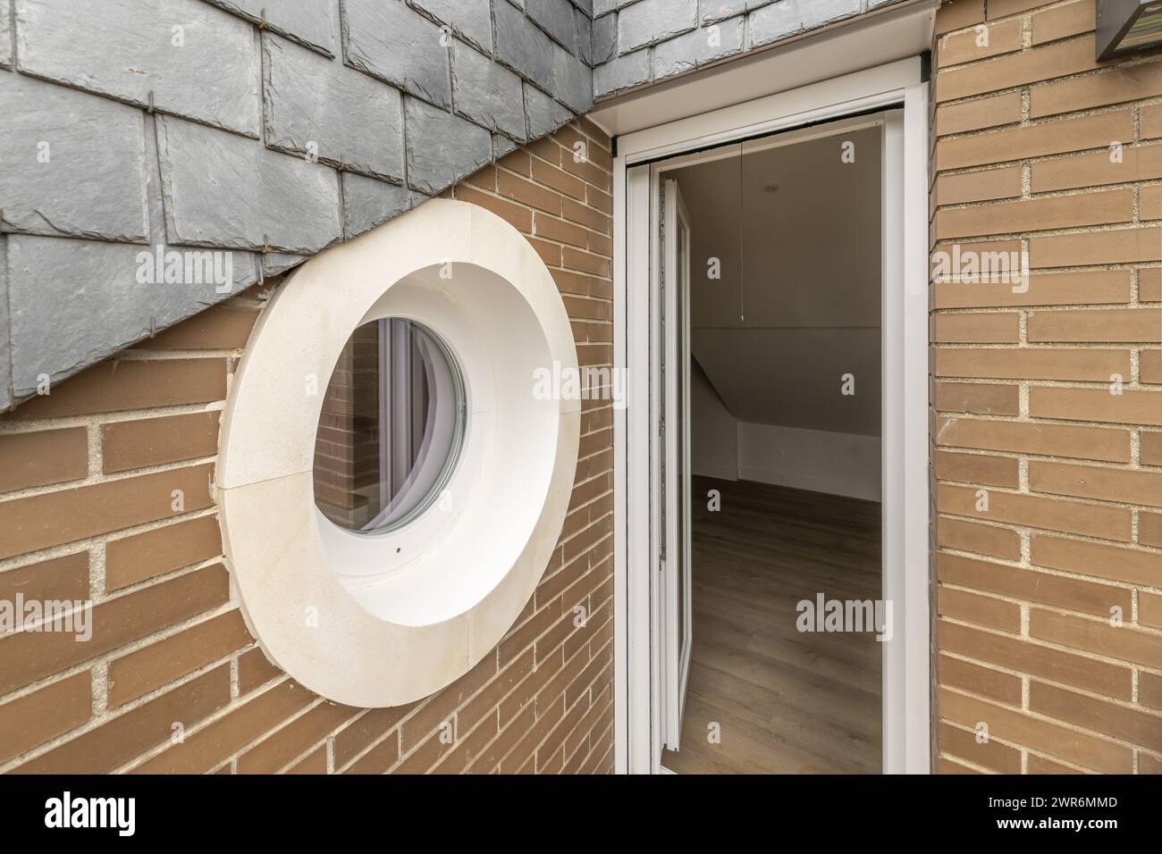 Kreisförmiges Fenster oder Bullauge einer kleinen Terrasse mit weißem Rahmen und braunen Ziegelwänden und weißer Aluminiumtür Stockfoto